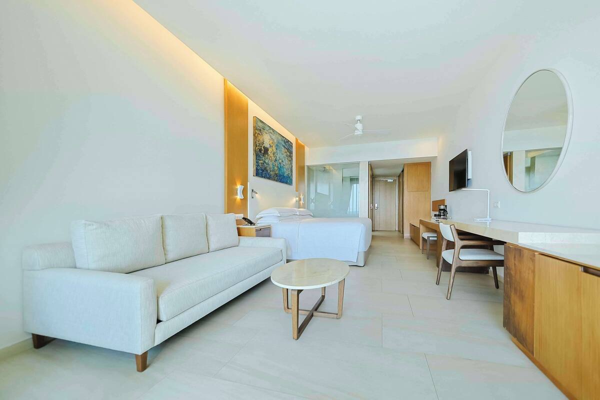 A modern hotel room with a sofa, a small round table, a bed, a desk with a chair, and a large mirror.