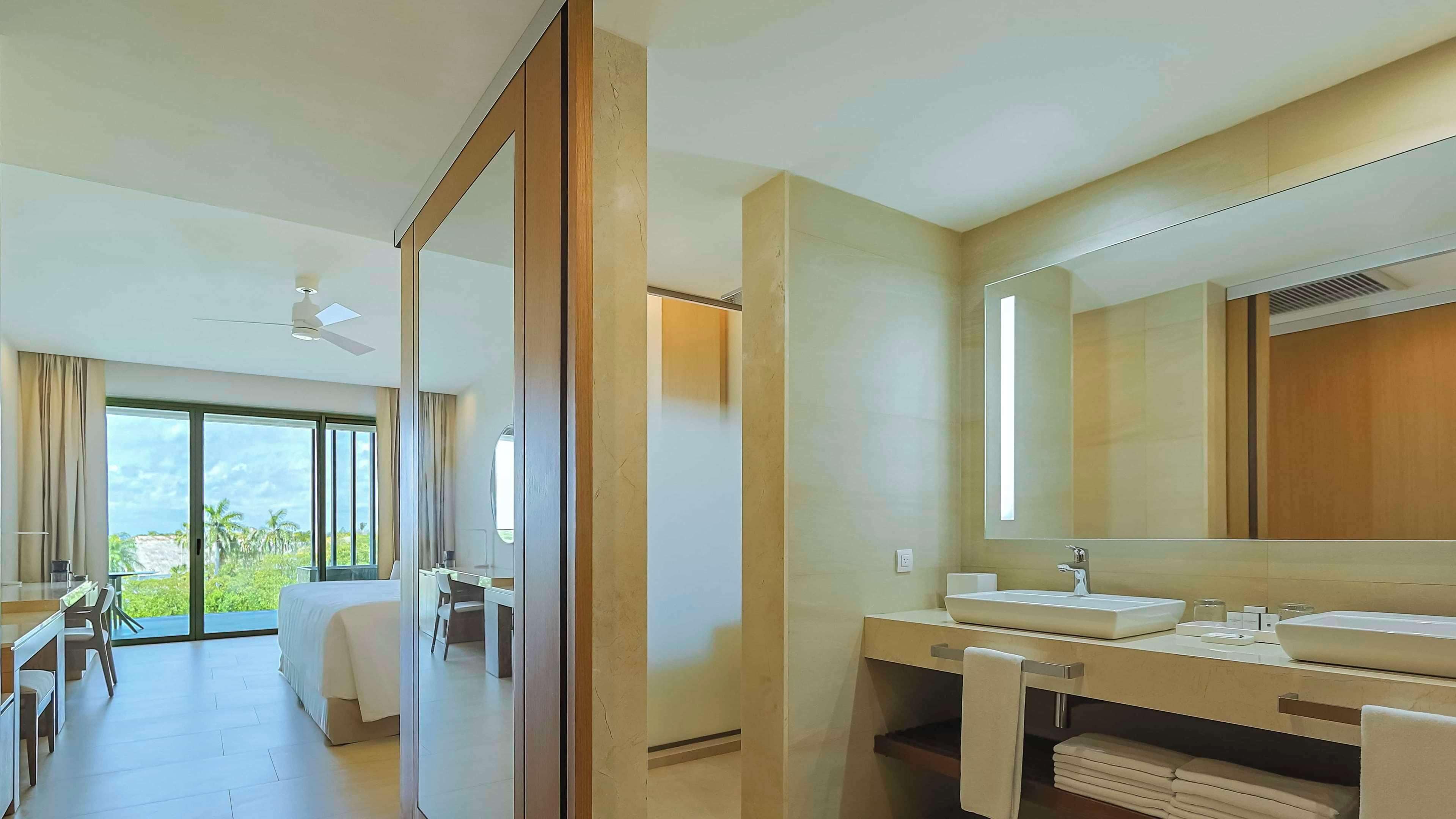 A modern hotel room with a large bed, a desk, a bathroom with a large mirror, and a view of greenery outside.