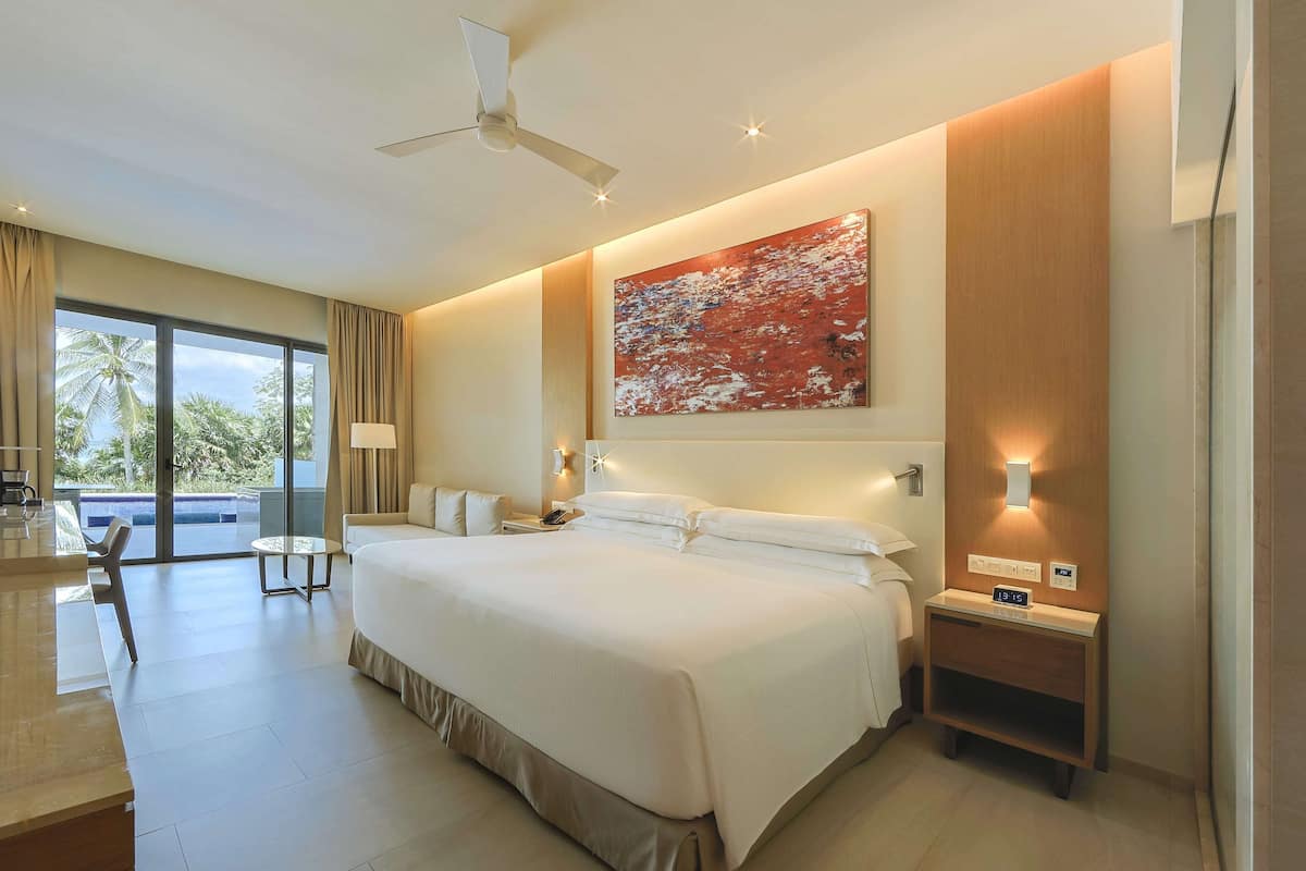 A modern hotel room with a large bed, a ceiling fan, a nightstand with a clock, and a view of palm trees through the window.