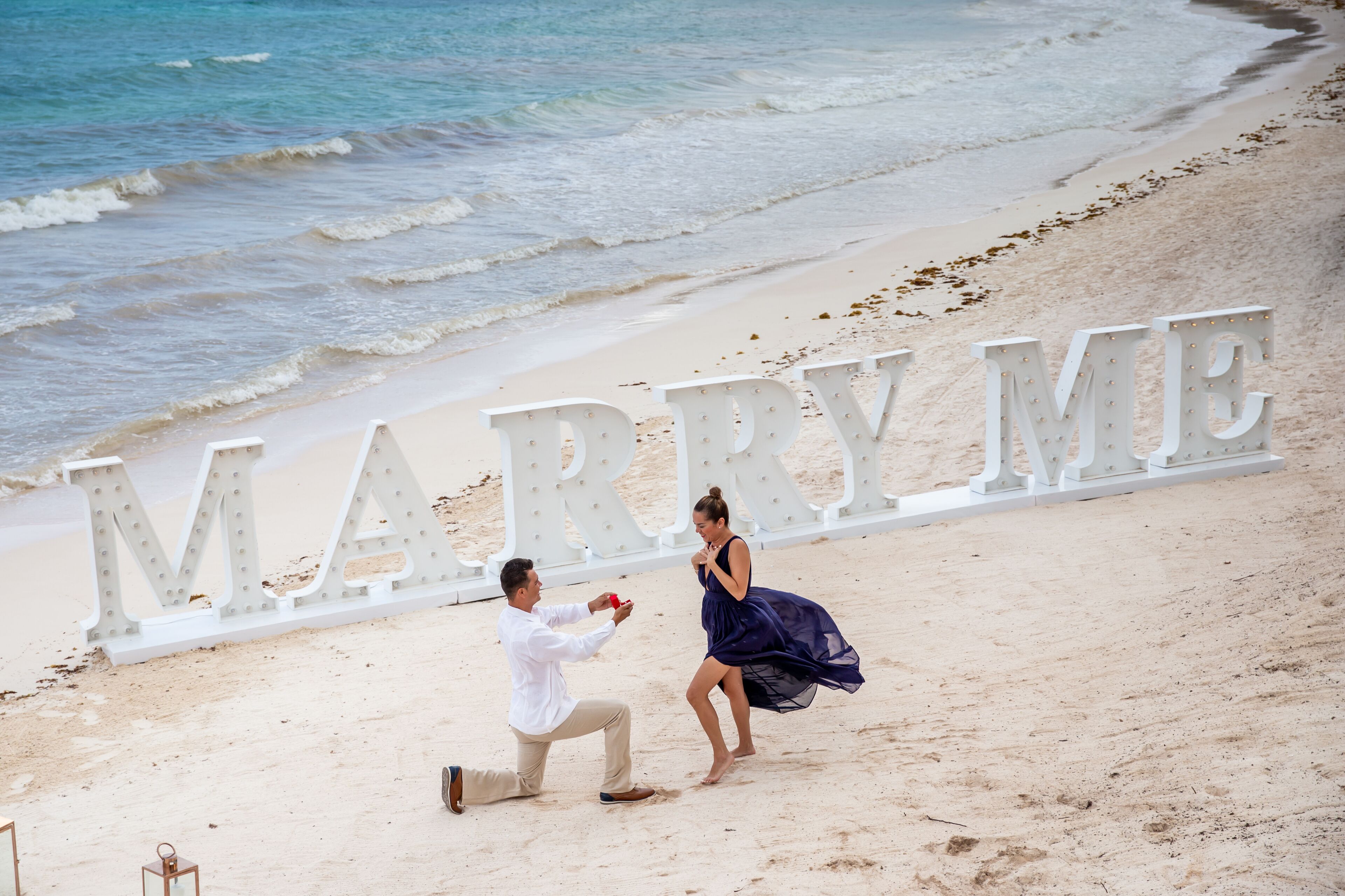 Ein Strandbild mit einem großen, beleuchteten Schild, auf dem 'MARRY ME' steht.