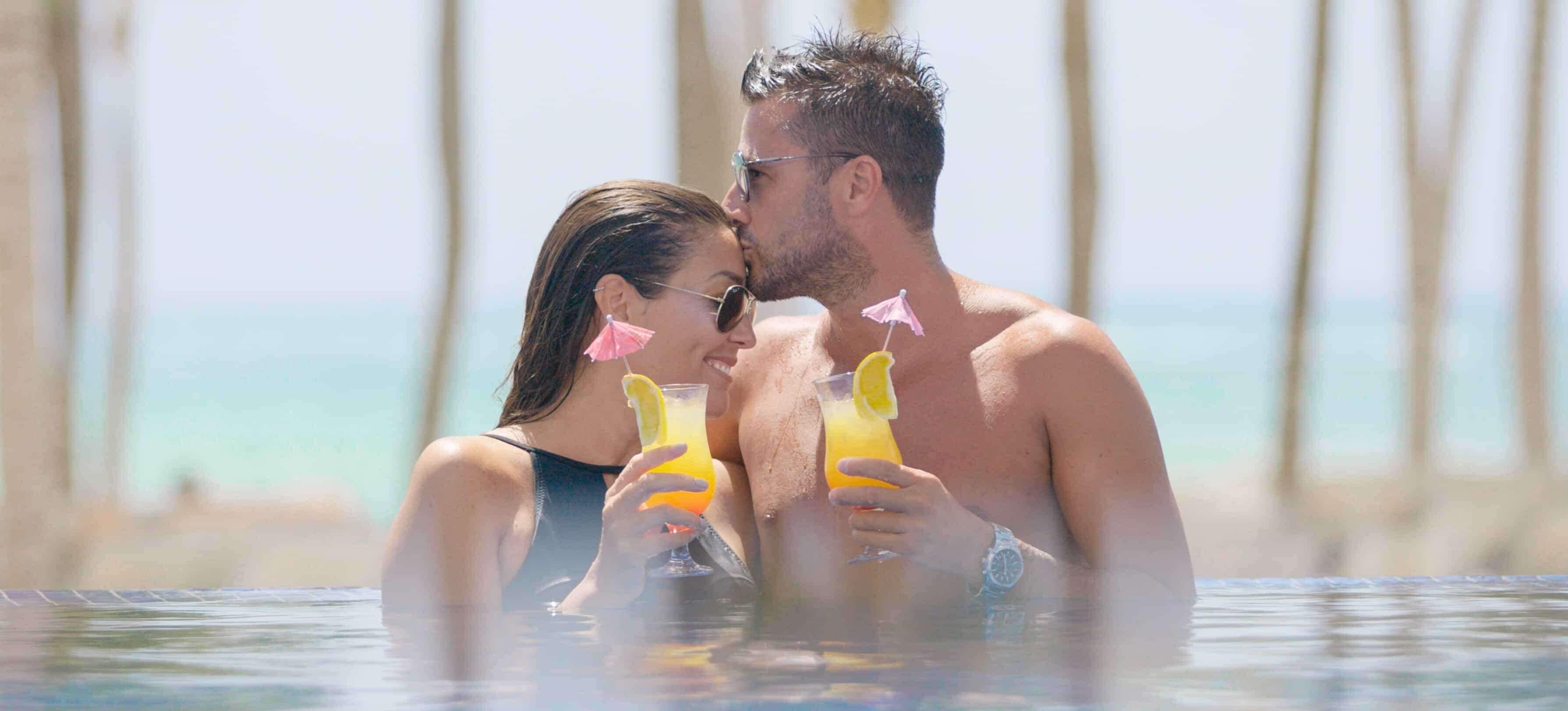 Una pareja en una piscina, con bebidas, rodeada de palmeras.