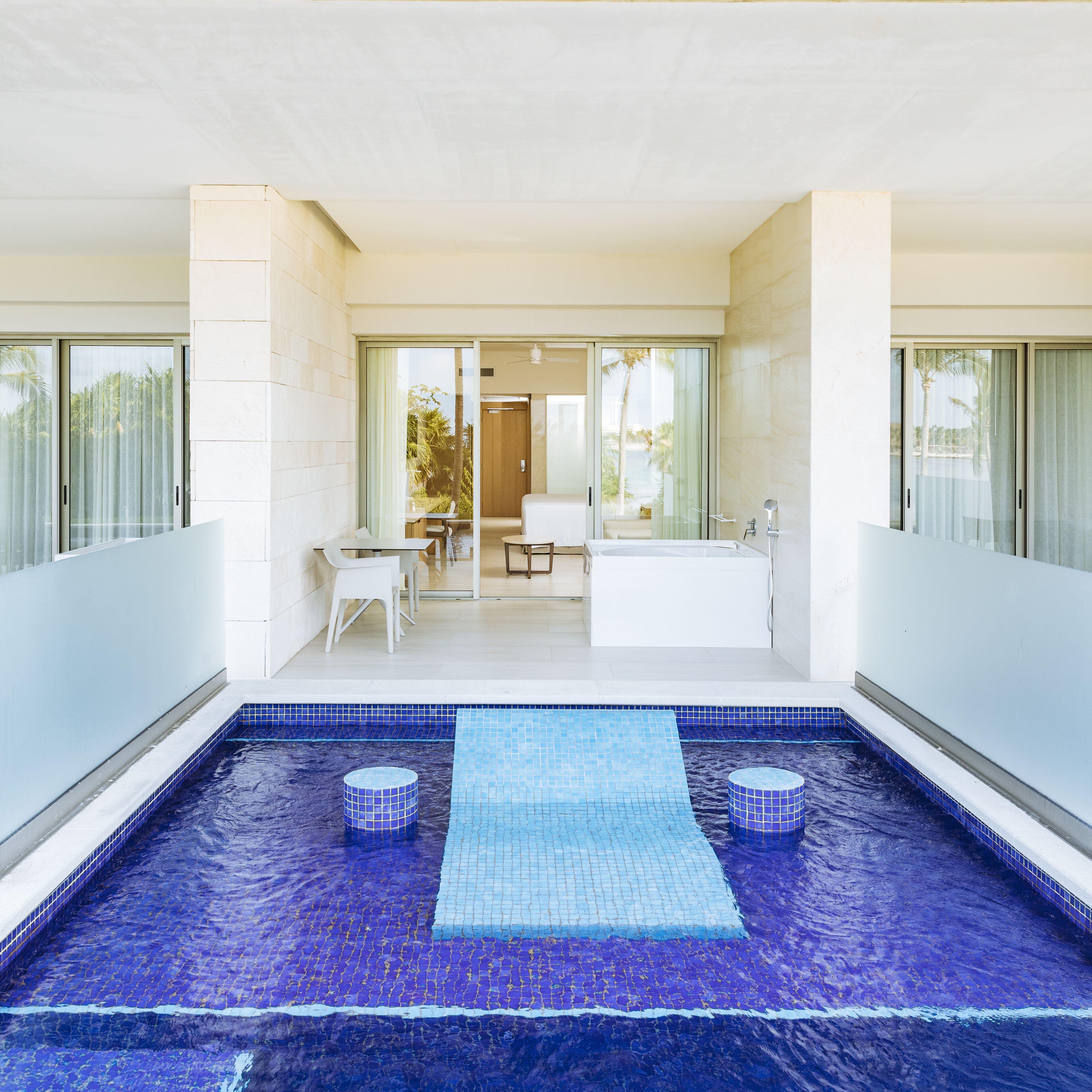 Un espace piscine intérieur moderne, doté d’un sol en mosaïque bleue, d’un coin salon avec une table et des chaises, et d’une grande baignoire.