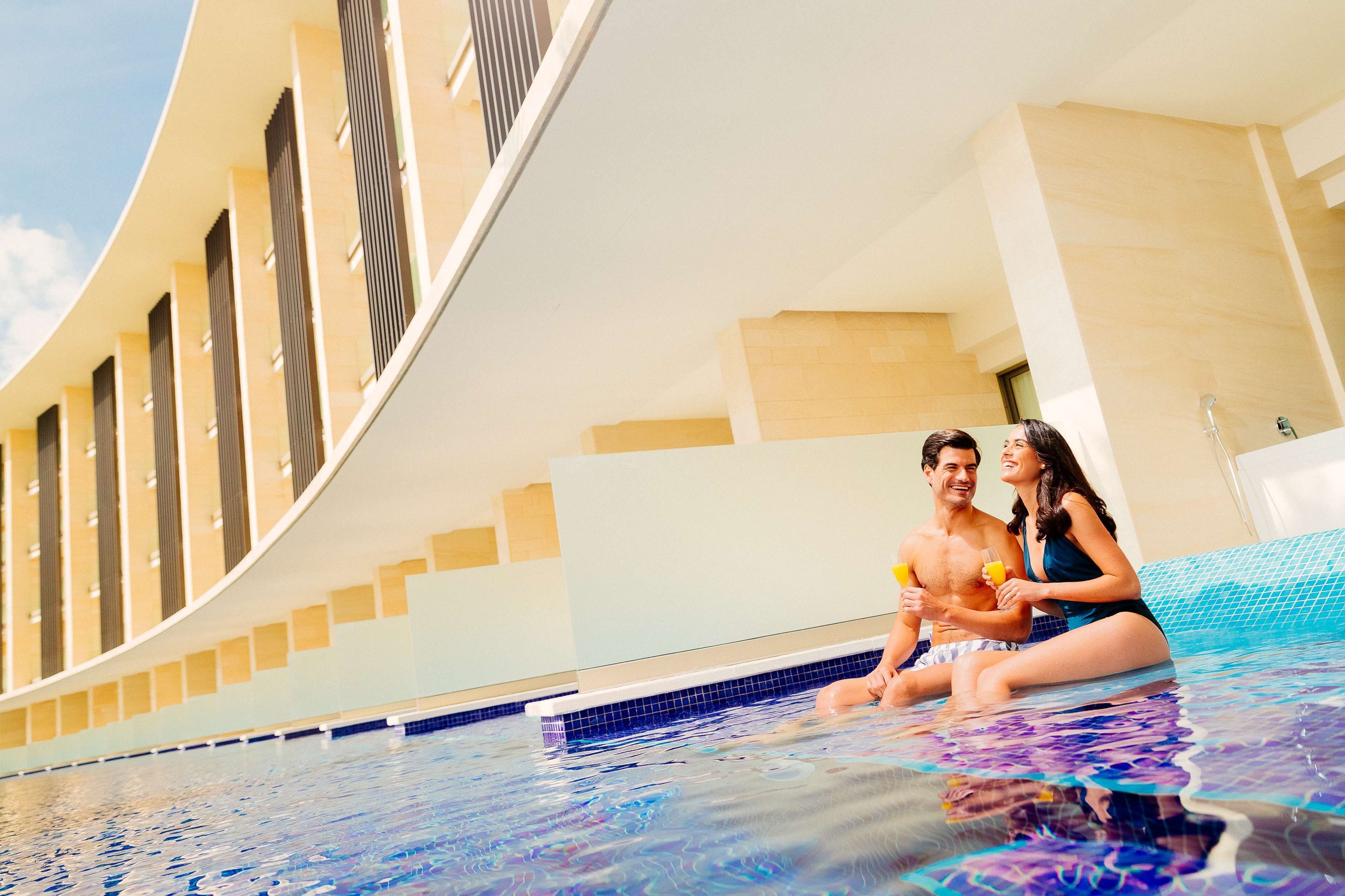 Una pareja sentada en una piscina interior moderna con un diseño arquitectónico curvo y vistas a un edificio con ventanales verticales.