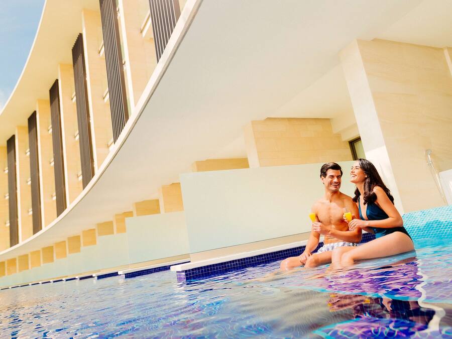 Un couple est assis dans une piscine intérieure moderne, dotée d’une conception architecturale aux lignes courbes et offrant une vue sur un bâtiment aux fenêtres verticales.