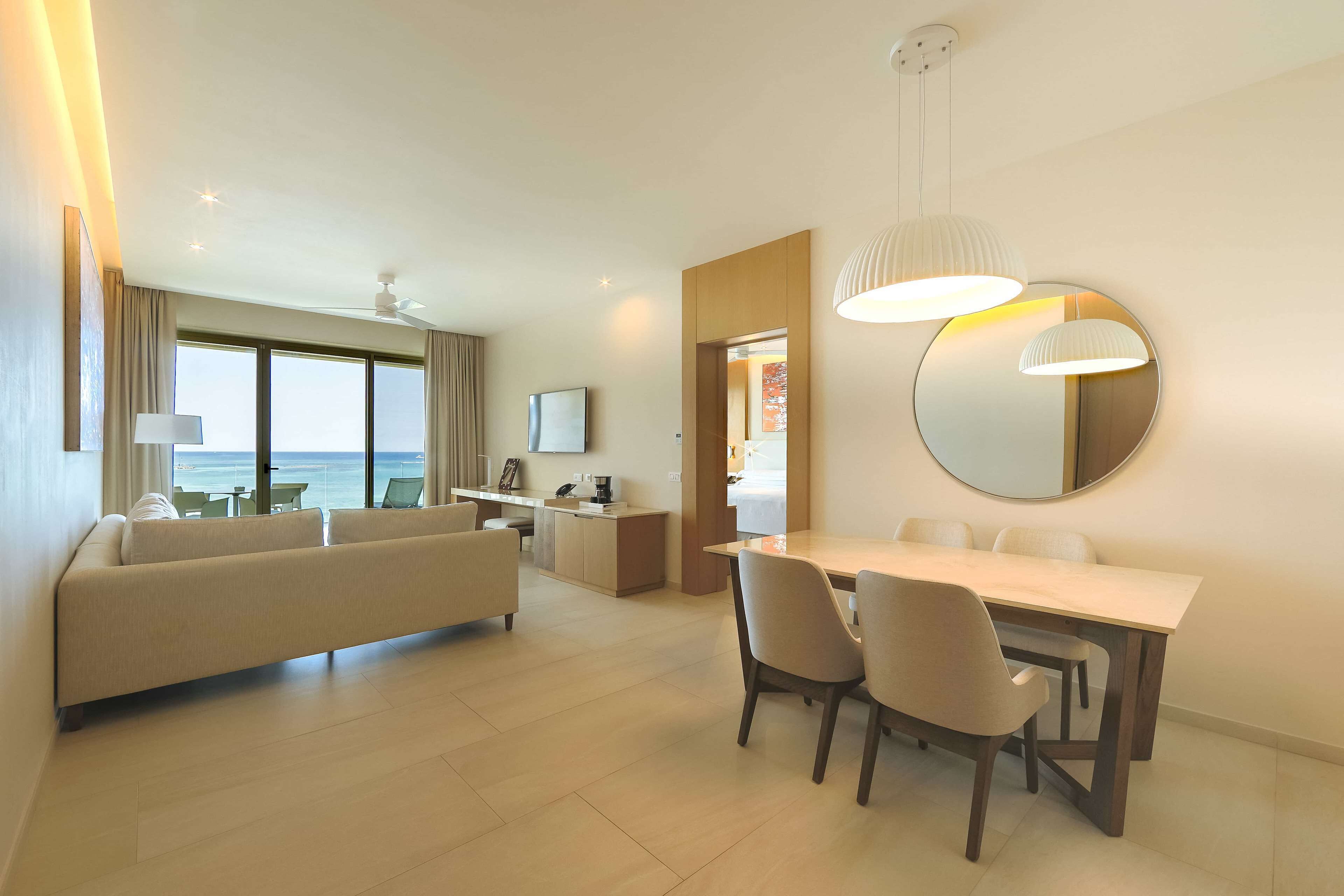 A modern hotel room with a dining area, a sofa, a desk, and a large window overlooking the sea.