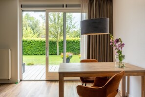 A room with a wooden table, two chairs, a floor lamp, and a view of a garden through a sliding glass door.