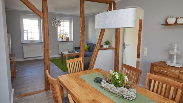 A dining area with a wooden table, chairs, a hanging light fixture, and a view into the living room. - Holiday home Eifelträume for 6 people (Birresborn)