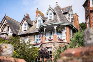 Traditional stone house with a gabled roof and multiple chimneys, featuring a bay window and balcony with hanging flower baskets amidst lush greenery.