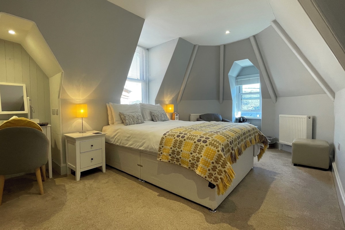 A bedroom with a sloped ceiling, a bed with a patterned blanket, bedside lamps, a chair, a desk with a computer, and a window.