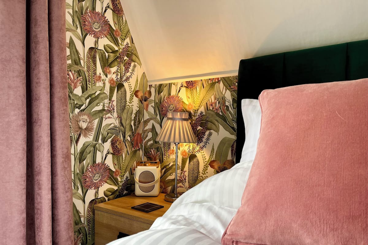 A neatly made bed with white and pink bedding, a bedside lamp, and a radio on a wooden nightstand. The wall features a floral wallpaper design.