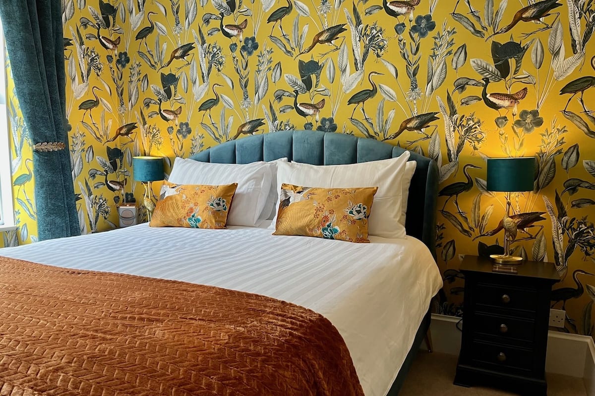 A bed with a white duvet and two pillows, a blue upholstered headboard, a brown woven throw, and a nightstand with a lamp.