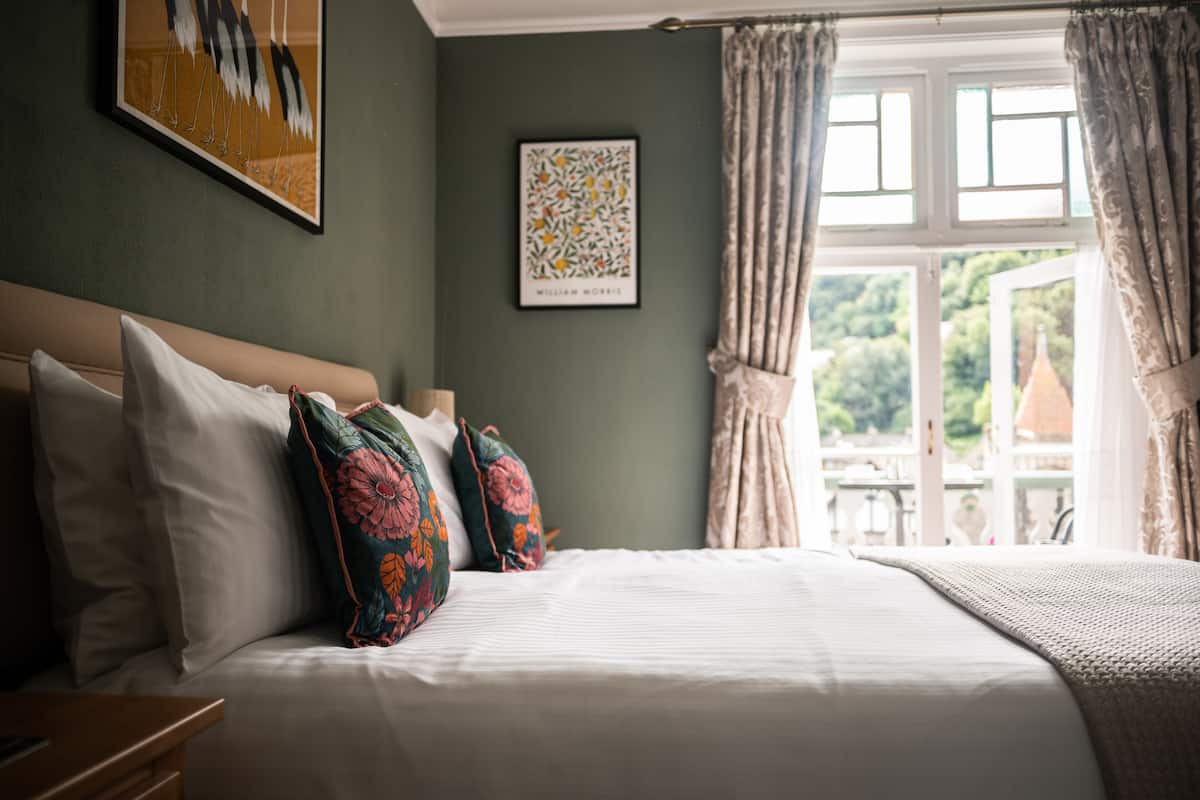A neatly made bed with white linens and colorful pillows, a framed artwork on the wall, and a window with curtains.
