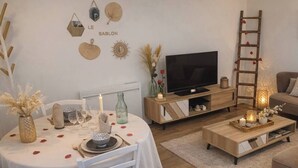 Una sala de estar acogedora con una mesa redonda para dos personas, un televisor sobre un mueble de madera y una escalera apoyada en la pared.