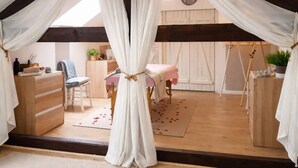 Un dormitorio acogedor en el ático, con techo inclinado, vigas de madera, una cama con dosel blanco, un escritorio y una silla.