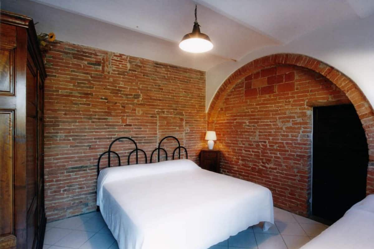 Una habitación con pared de ladrillo, una cama con ropa de cama blanca, cabecero metálico, mesita de noche pequeña con una lámpara y un armario de madera.