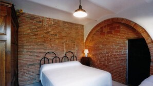 A room with a brick wall, a bed with white bedding, a metal headboard, a small bedside table with a lamp, and a wooden wardrobe. - Podere Castagno (Lorenzana)
