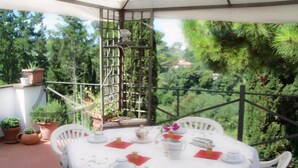 A round table with a white tablecloth, surrounded by white chairs, set on a balcony with potted plants and a view of greenery. - Podere Castagno (Lorenzana)