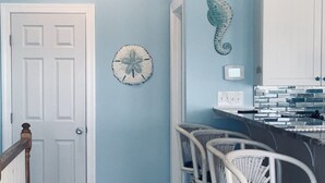 Un couloir avec une porte blanche, une enseigne fixée au mur, un hippocampe décoratif et des chaises en osier.