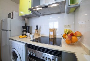 A kitchen with a washing machine, a coffee maker, a bowl of fruit, and a microwave.