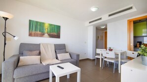 A living room with a sofa, a coffee table, a painting, a lamp, and a kitchen area with a dining table and chairs. - Borumbot 8.3.27 VistaMar (Calpe)