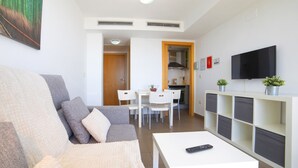 A living room with a sofa, a coffee table, a TV, and a kitchen area with chairs and a table. - Borumbot 8.3.27 VistaMar (Calpe)