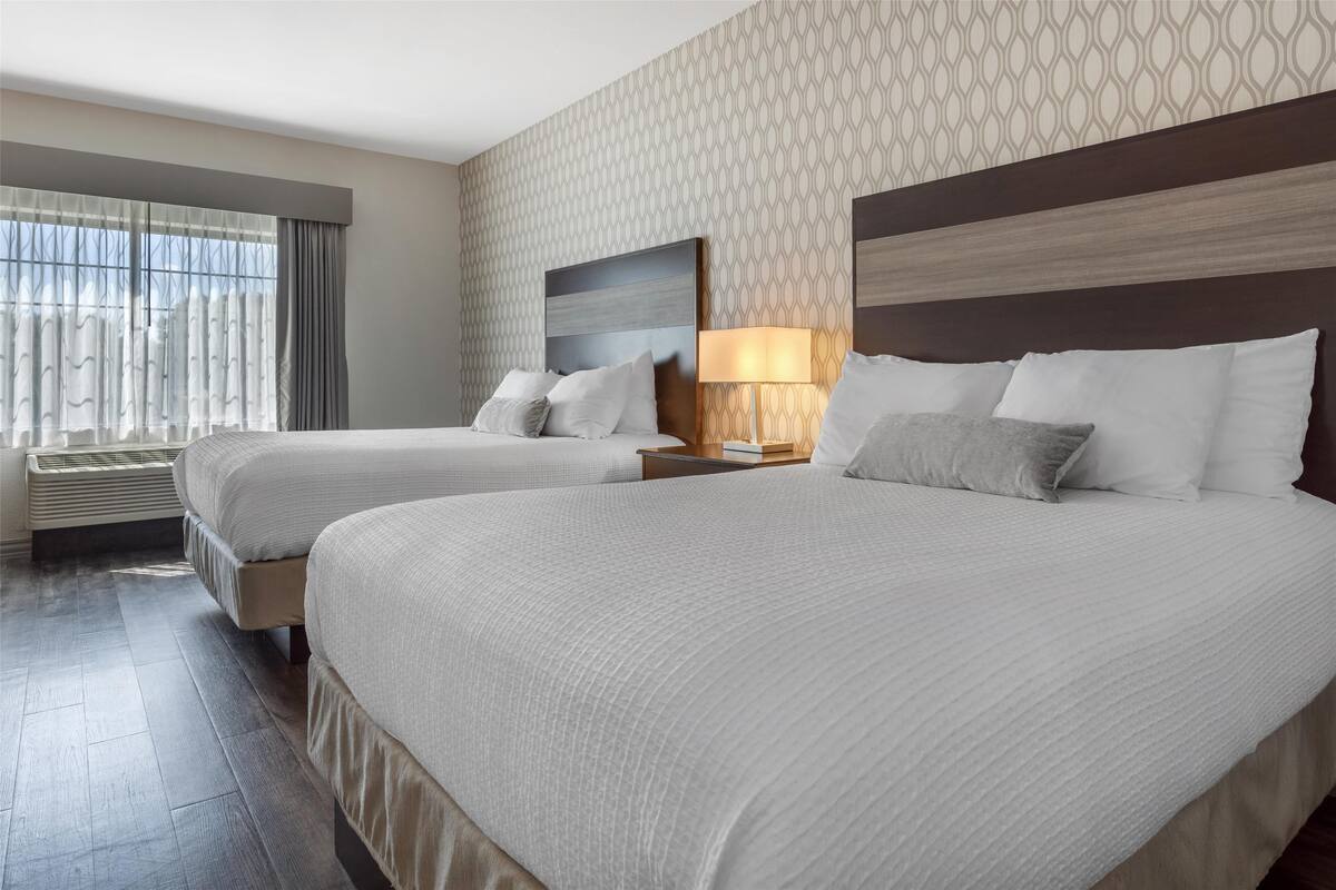 A hotel room with two beds, a large window with curtains, a patterned wallpaper, and a modern headboard.