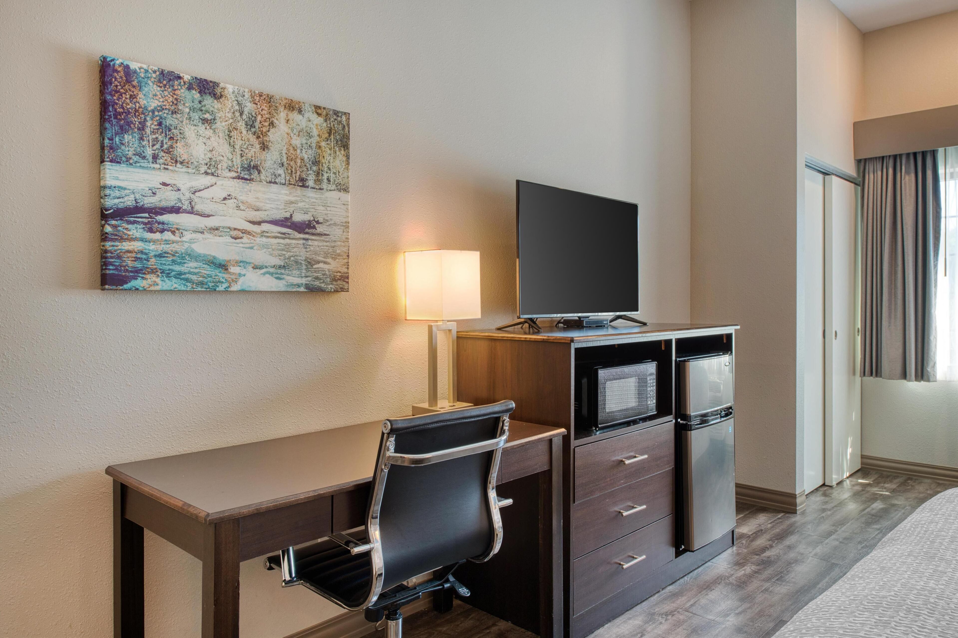 A hotel room with a desk, a flat-screen TV, a refrigerator, a microwave, a lamp, and a painting on the wall.