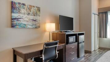 A hotel room with a desk, a flat-screen TV, a refrigerator, a microwave, a lamp, and a painting on the wall.