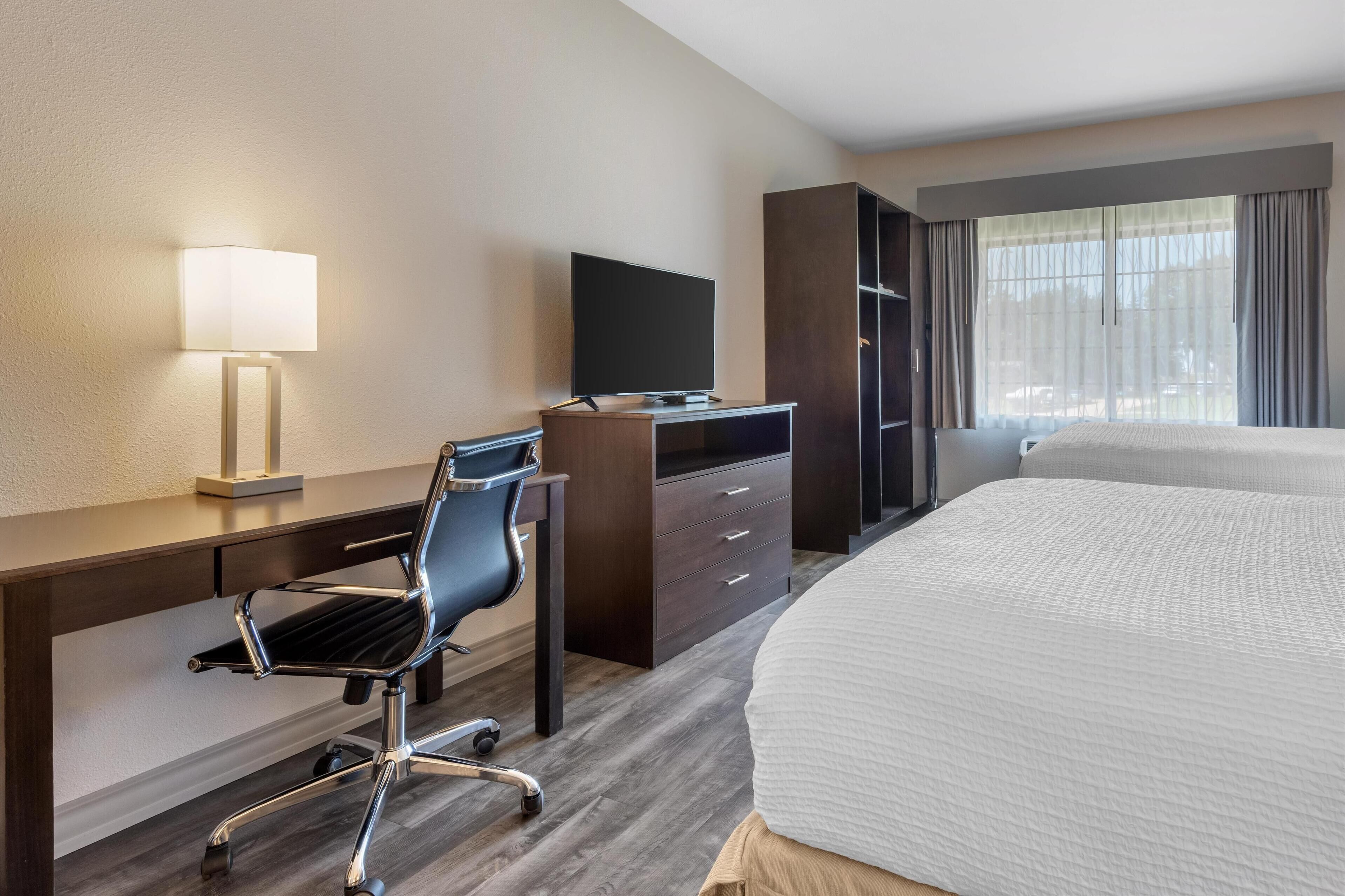 Una habitación de hotel con una cama, un escritorio con computadora y lámpara, una silla y un televisor.