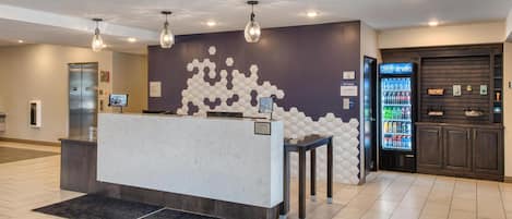 Uma área de recepção com balcão de mármore, um padrão hexagonal na parede, um dispensador de bebidas e uma tela digital.