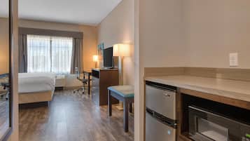 A hotel room with a bed, desk, chair, lamp, microwave, and a small kitchenette.