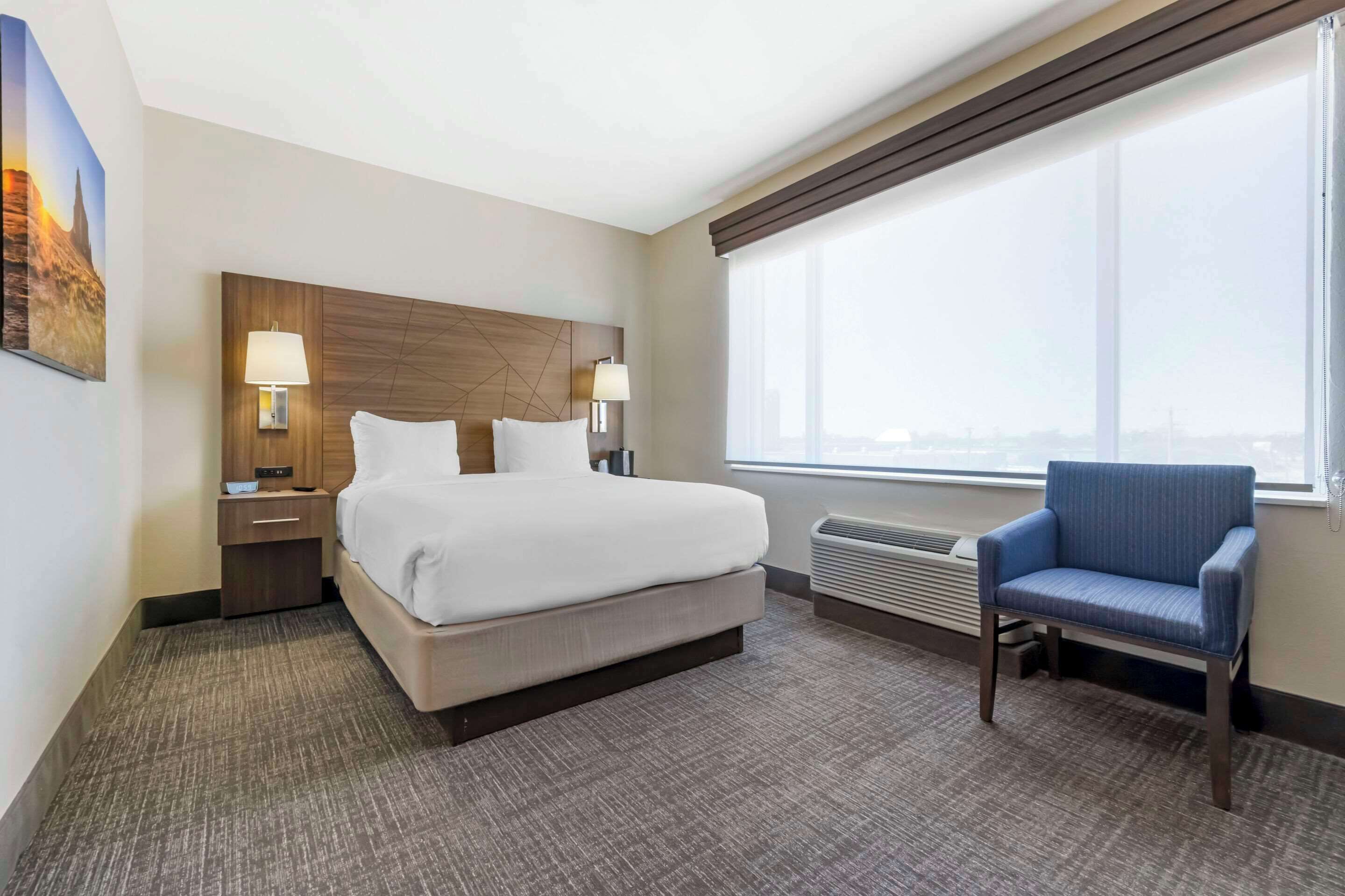 A hotel room with a bed, two bedside tables with lamps, a chair, a large window, and a wall-mounted air conditioner.
