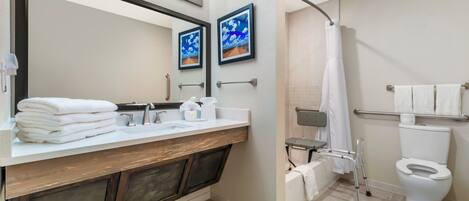 A modern bathroom with a large mirror, two framed pictures, a sink with a towel stack, a shower with a grab bar, and a toilet with a towel rack.
