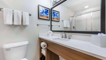 A modern bathroom with a white toilet, a wooden vanity with a sink, and a large mirror. There are towels hanging on a rack and a framed picture on the wall.