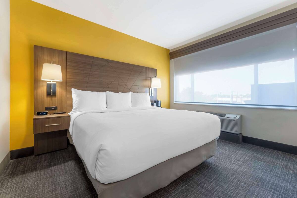 A hotel room with a large bed, two bedside tables with lamps, a window with a view, and a thermostat on the wall.