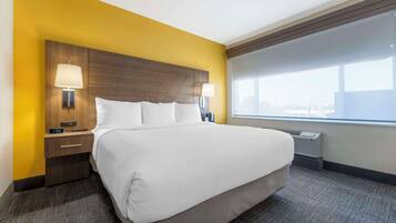 A hotel room with a large bed, two bedside tables with lamps, a window with a view, and a thermostat on the wall.