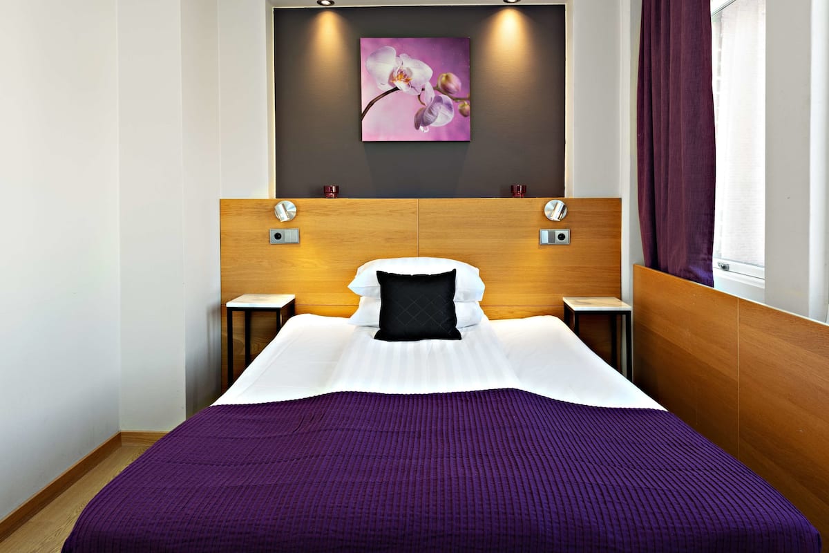 A modern hotel room with a large bed, wooden headboard, two bedside tables, and a wall-mounted artwork of an orchid.