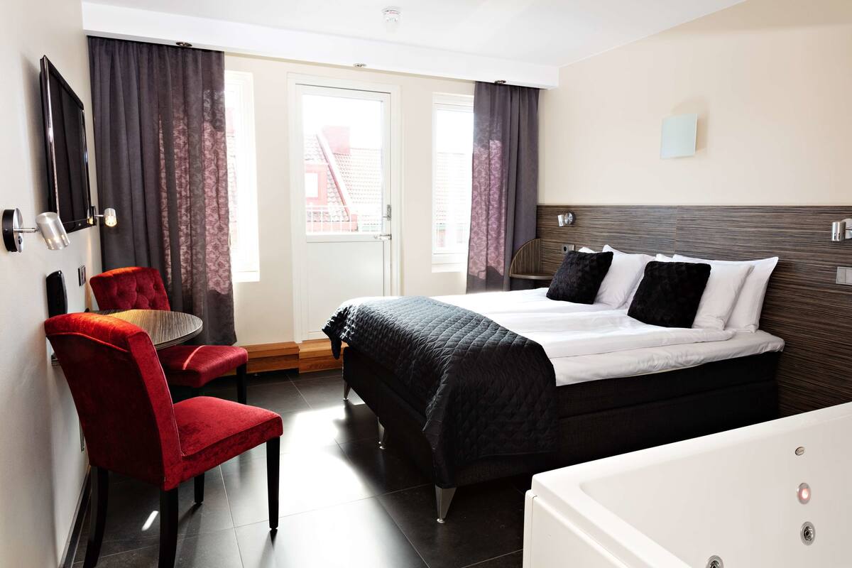 A hotel room with a bed, a red chair, a small table, a TV, and a window with curtains.