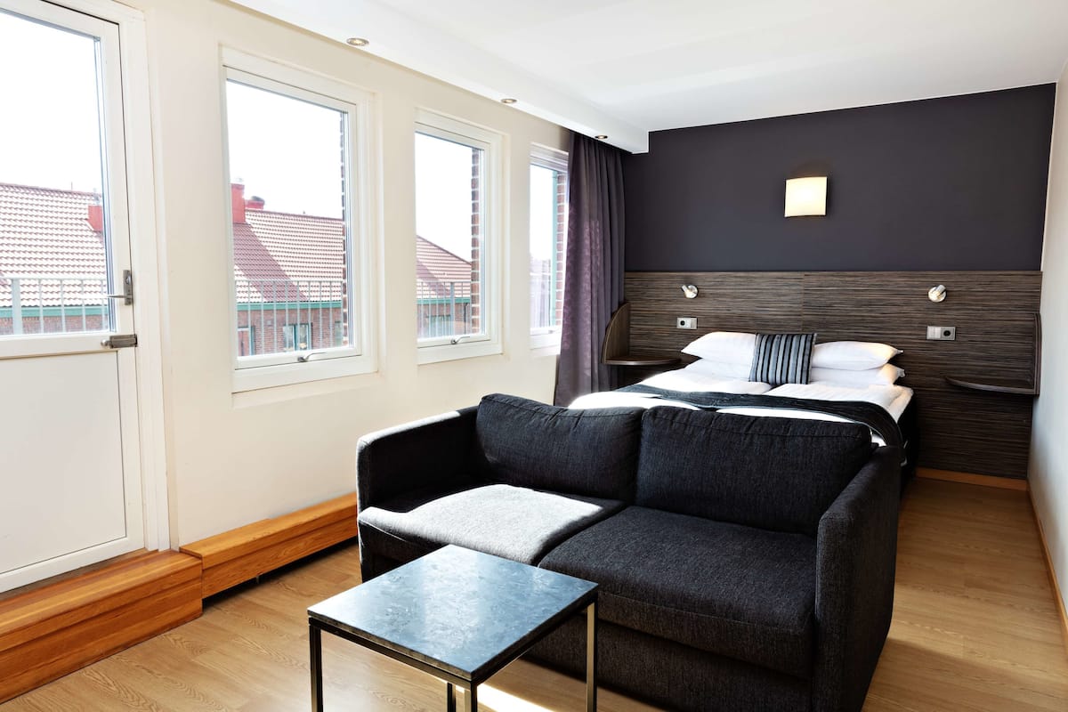A modern hotel room with a bed, a sofa, a small table, and large windows offering a view of buildings outside.
