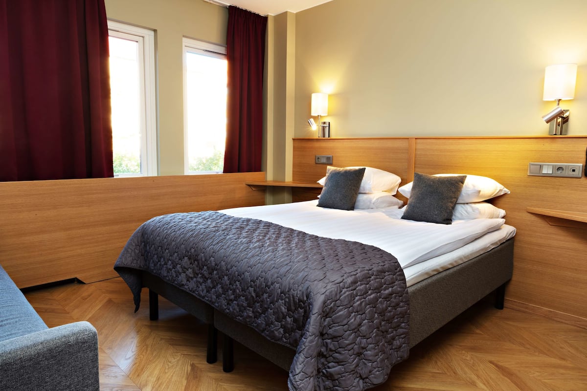 A hotel room with a large bed, wooden headboard, two bedside lamps, and a window with curtains.