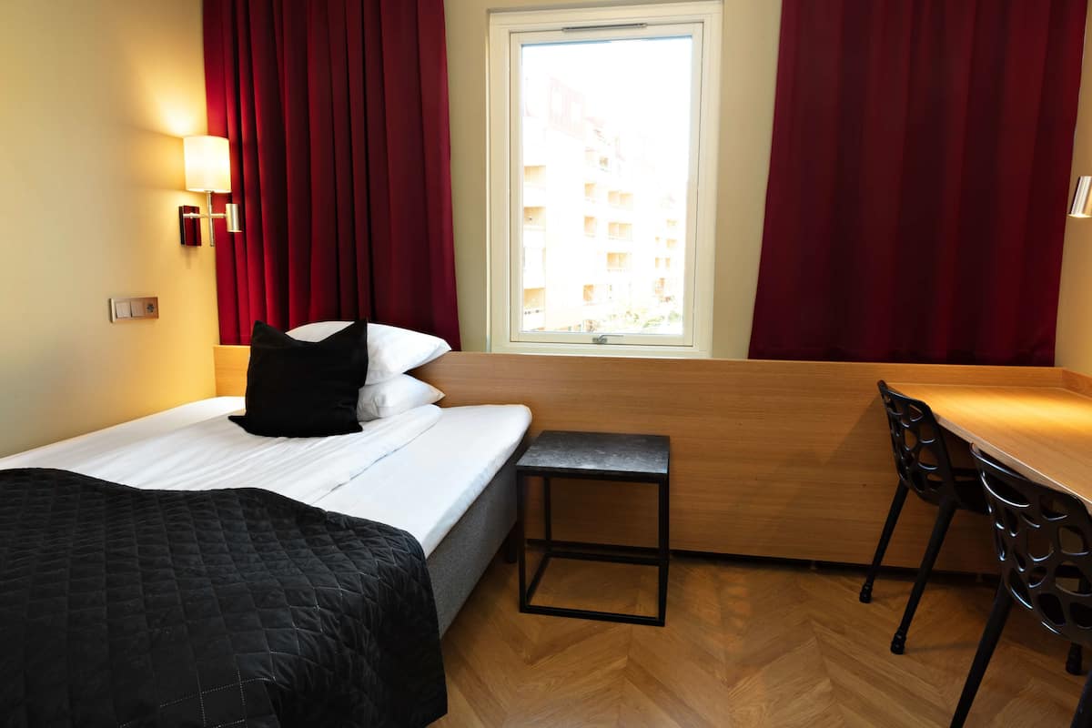 A hotel room with a bed, a small table, two chairs, a window with curtains, and a lamp on the wall.
