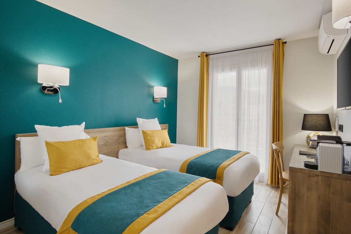A hotel room with two beds, teal walls, white bedding, and a desk with a lamp.