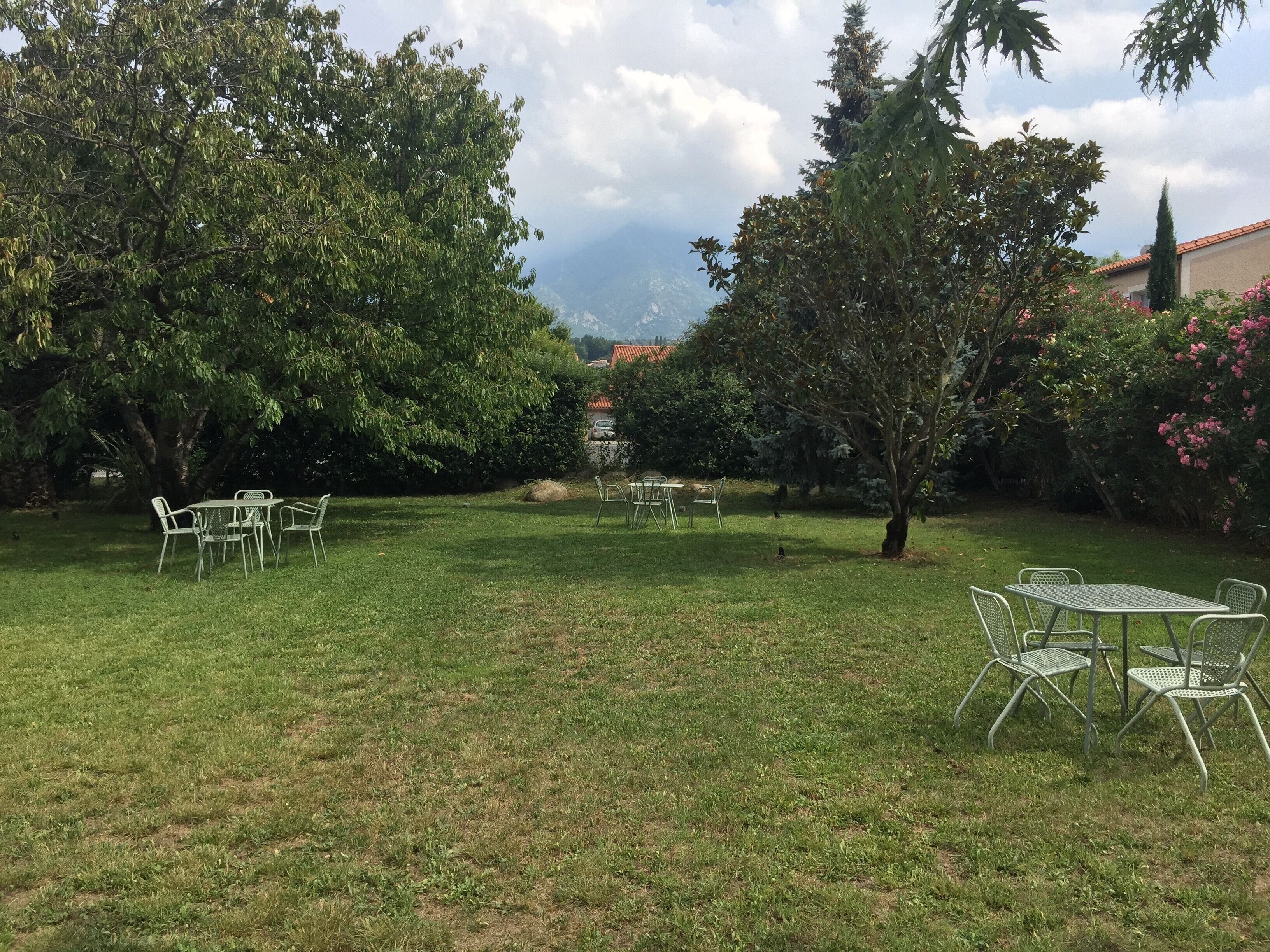A grassy outdoor area with metal tables and chairs, surrounded by trees and shrubs.