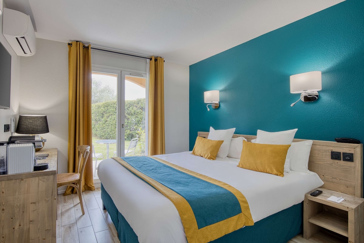 A hotel room with a large bed, a desk with a computer, and a view of greenery outside.