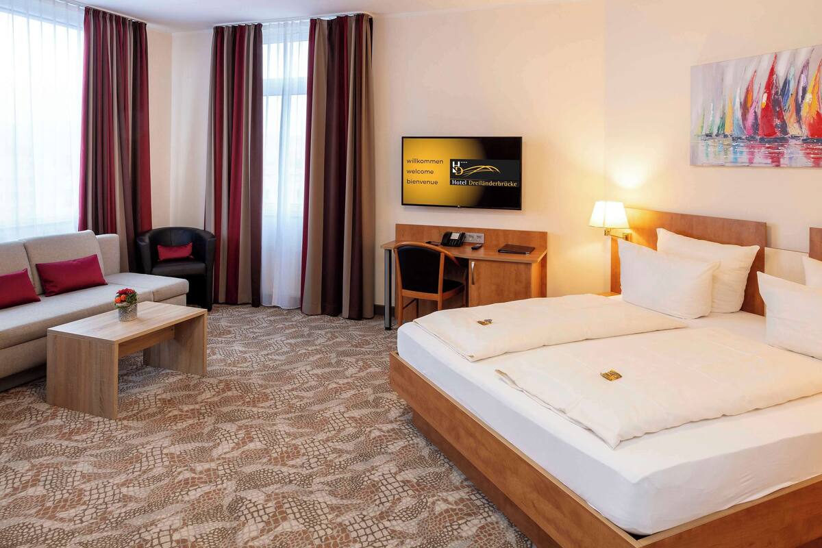 Habitación de hotel con cama, escritorio, sillón, televisión y un cuadro en la pared.