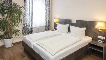 A double bed with white linens, two bedside lamps, a small bedside table with a clock, and a potted plant.