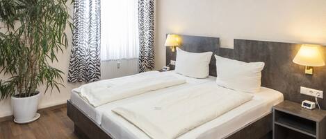 A double bed with white linens, two bedside lamps, a small bedside table with a clock, and a potted plant.