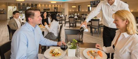 A restaurant setting with diners seated at tables, a waiter serving wine, and a bar area in the background.