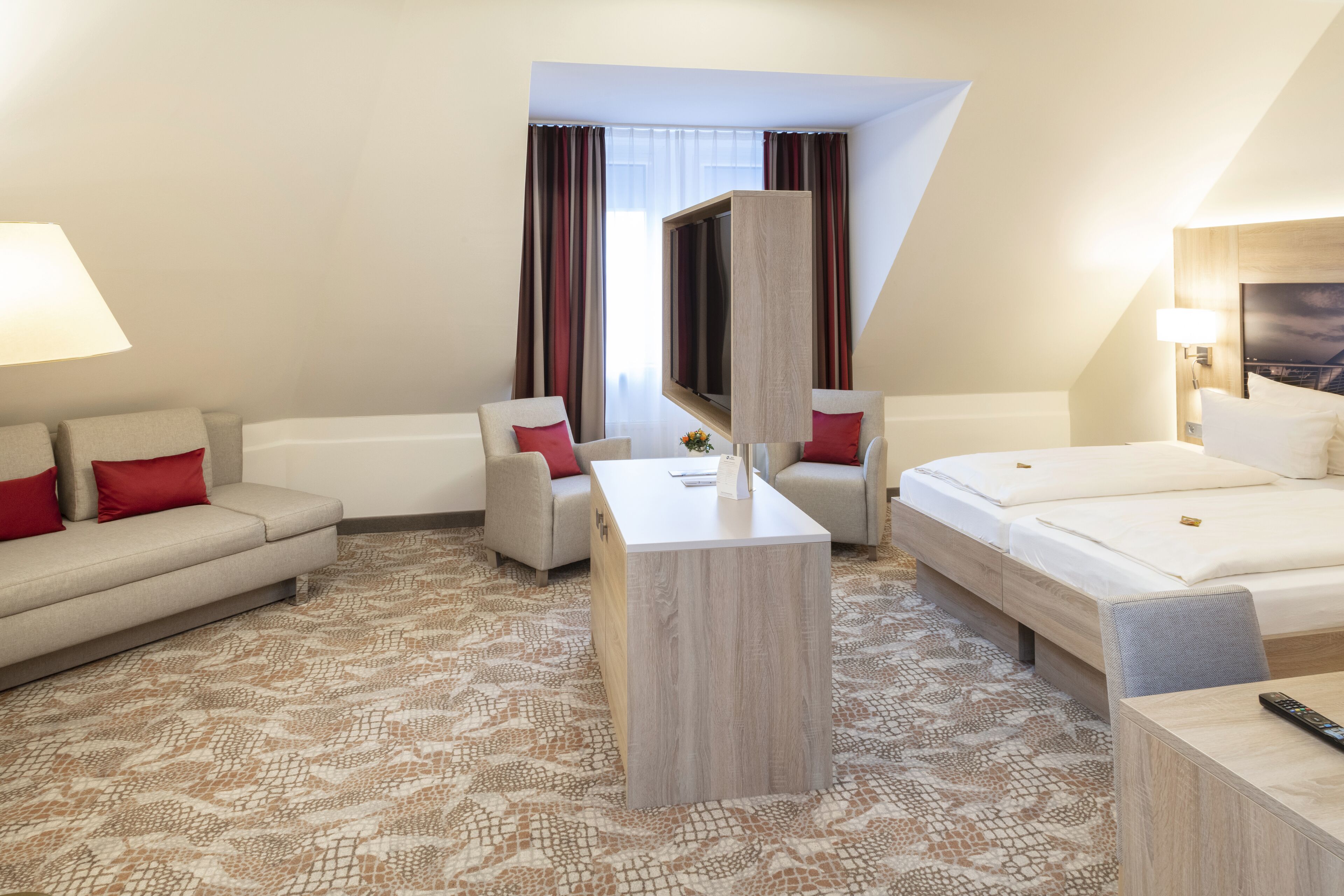 Ein Hotelzimmer mit einer Sitzecke, zwei Sesseln, einem Bett, einem Fernseher und einem Schreibtisch.