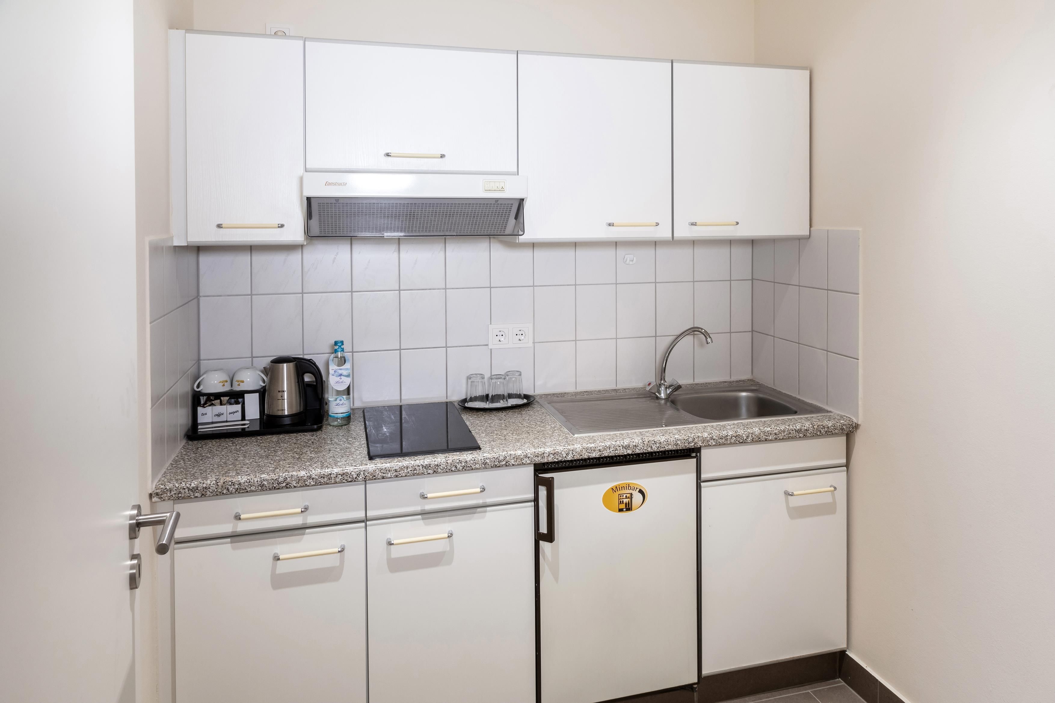 Cocina compacta con gabinetes blancos, encimera de granito, campana extractora integrada, lavamanos y cafetera.